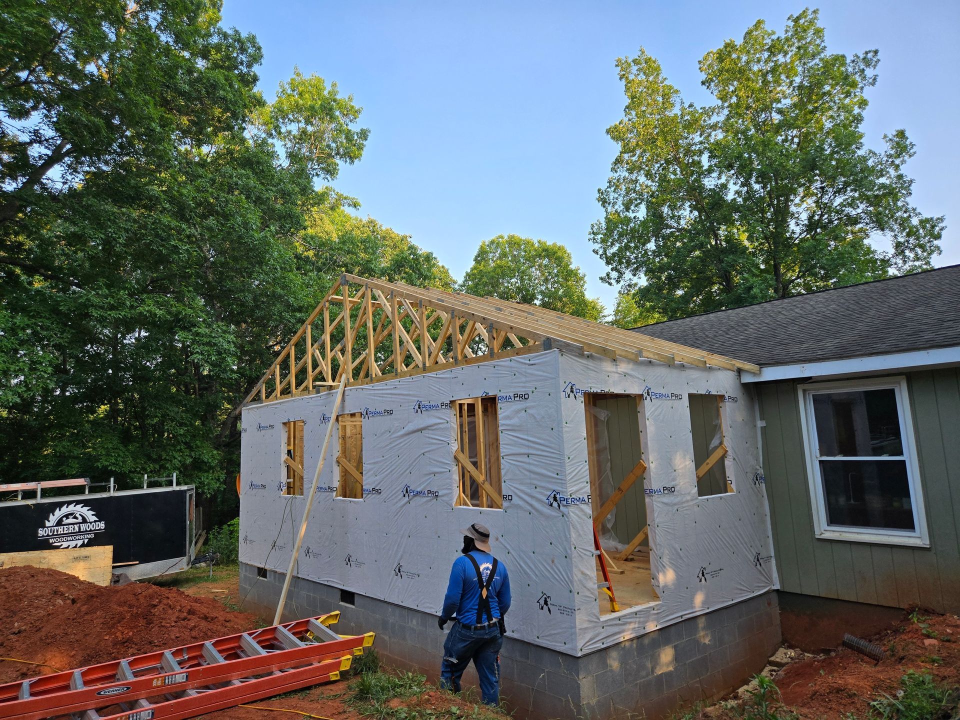 A person in work gear stands before a new room addition under construction, featuring exposed wooden trusses and wrap.
