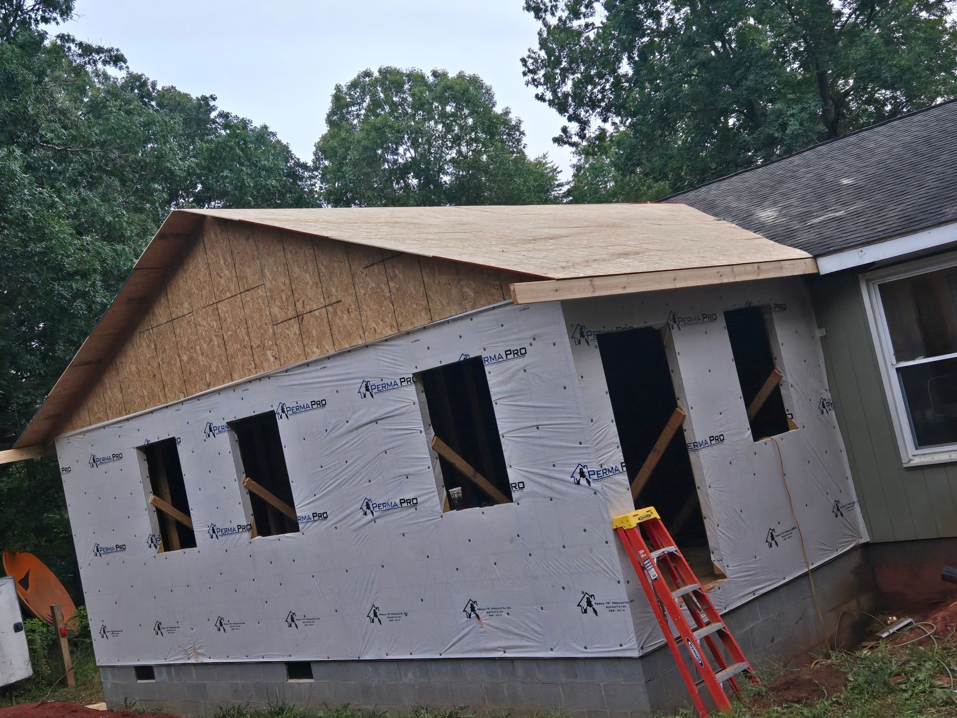 An unfinished house addition with plywood roofing, white weather-resistant wrap on the walls, and a red ladder nearby.
