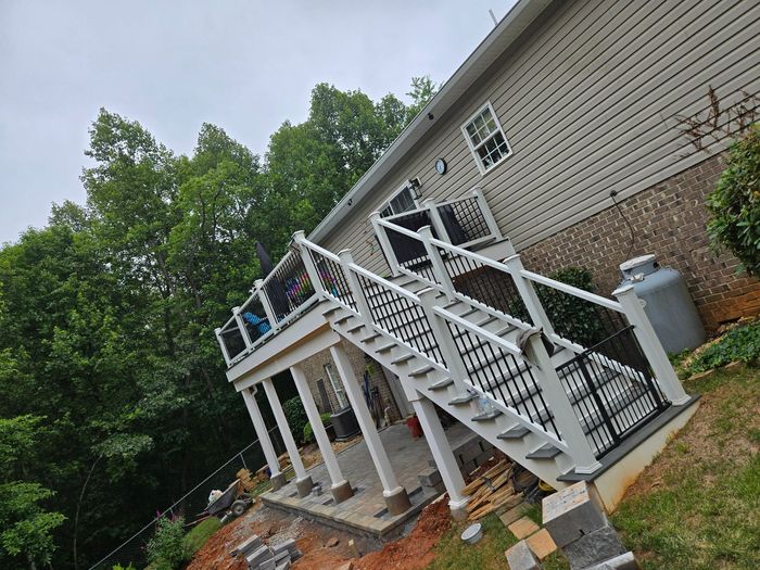 A raised wooden deck with white railings and stairs, leading down to a concrete patio area near a house with beige siding.