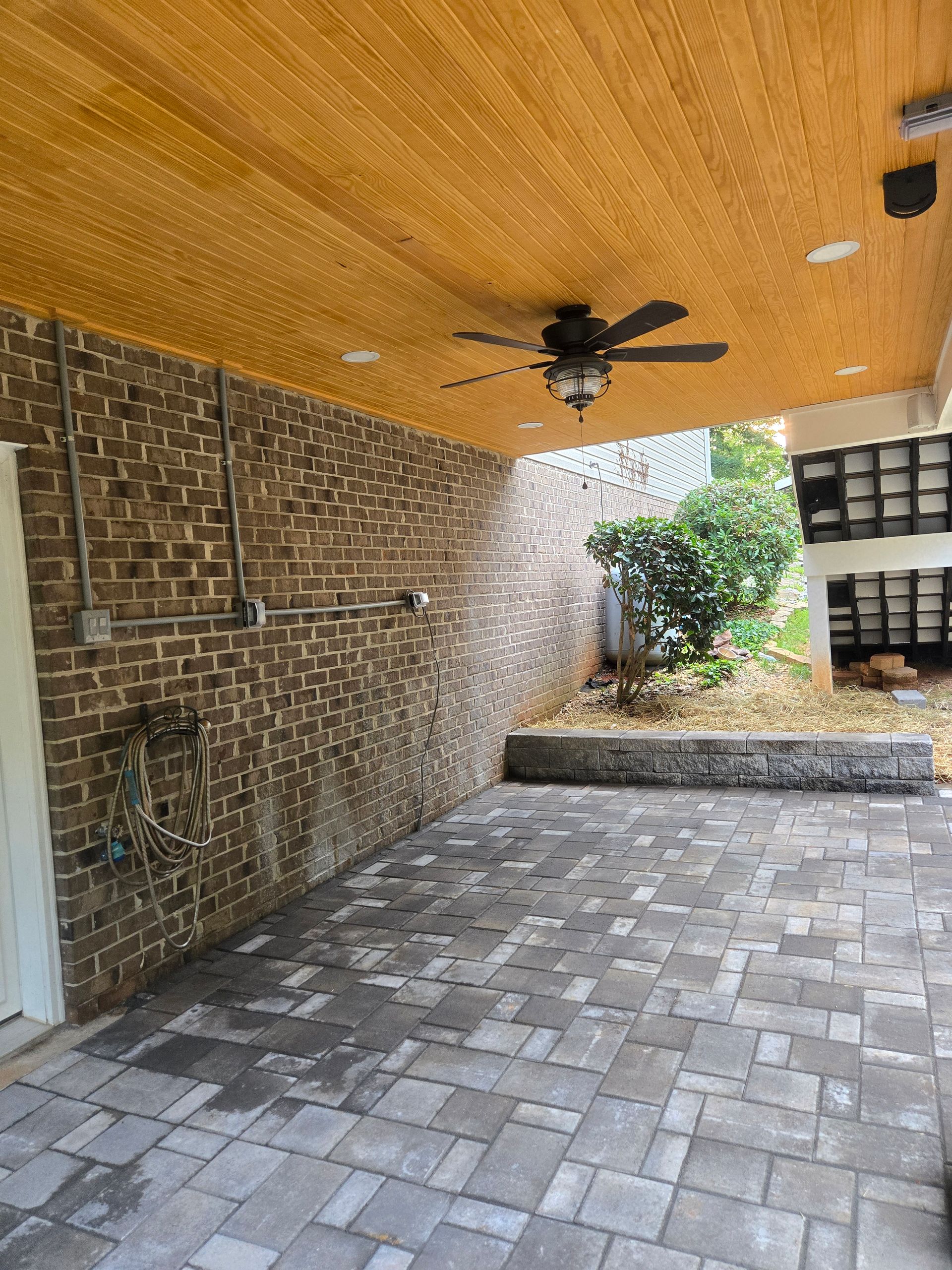 A covered patio with a wooden ceiling, a ceiling fan, a brick wall, and gray stone paver flooring.