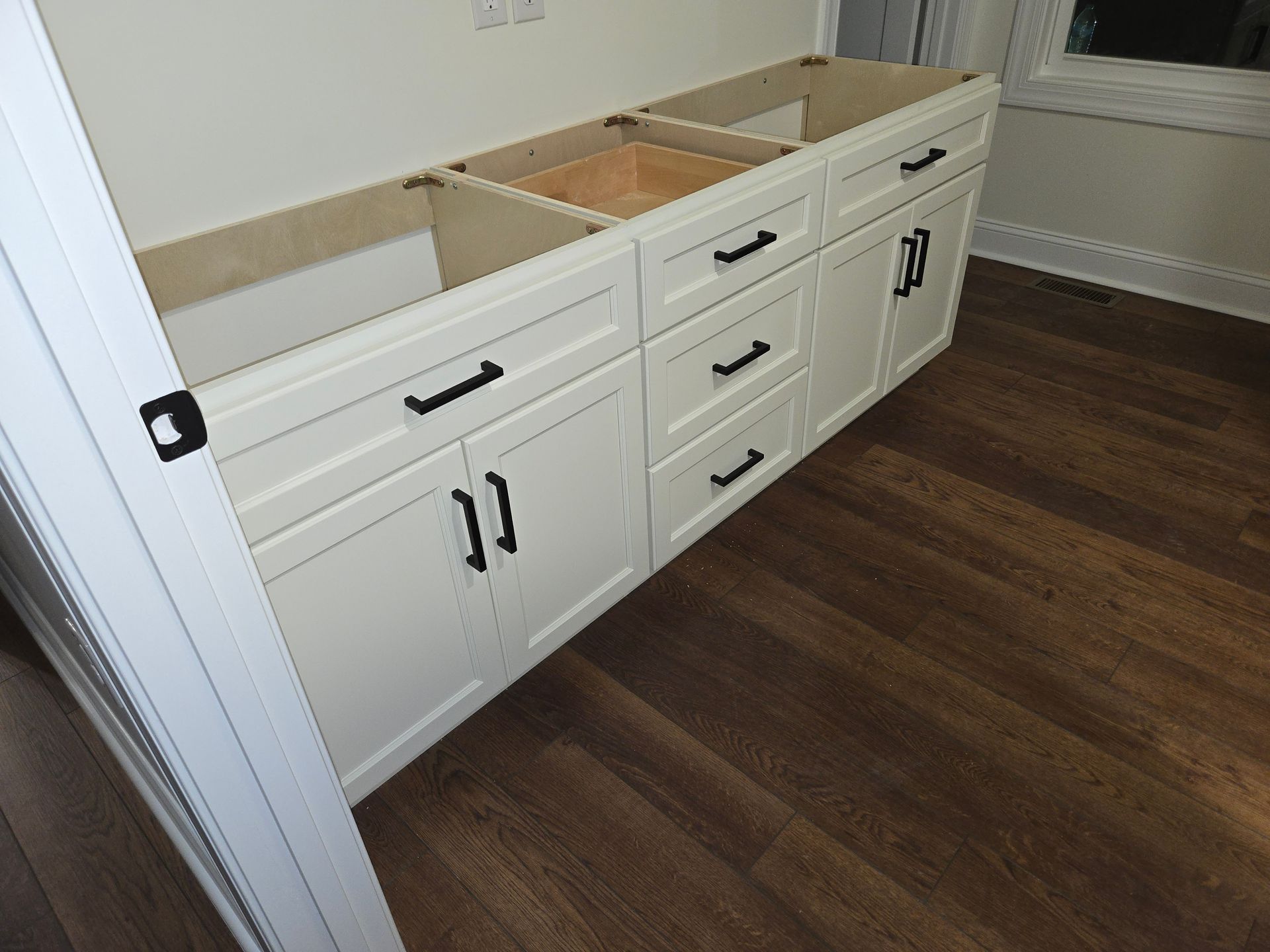 White bathroom vanity cabinets with black hardware and a wooden floor, viewed from an angle in an unfinished room.