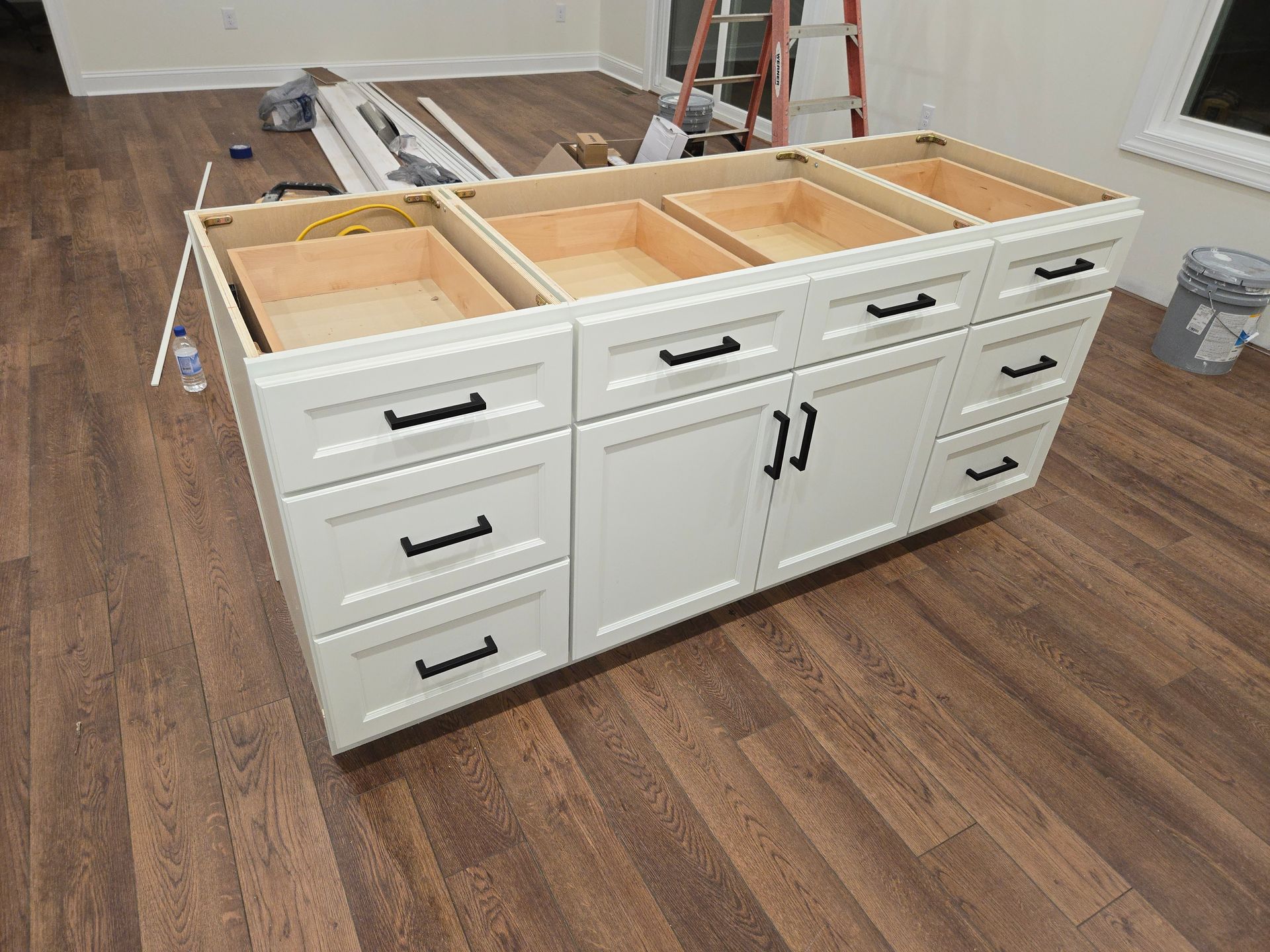 A white kitchen island cabinet with black hardware and open drawers, sitting on a wood-look floor in a room under renovation.