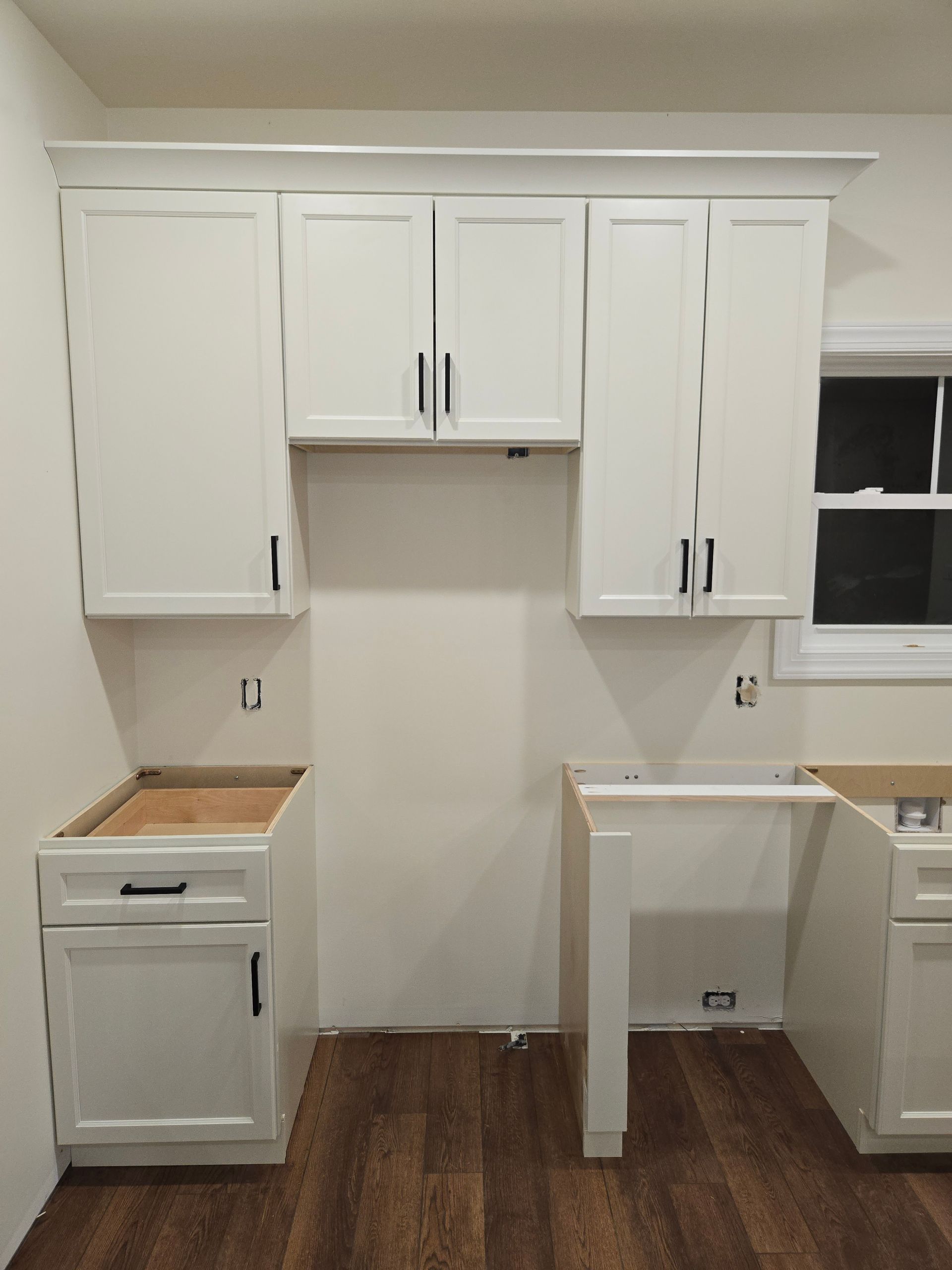 White kitchen cabinets installed against a wall with an open space for appliances and wood-look flooring.
