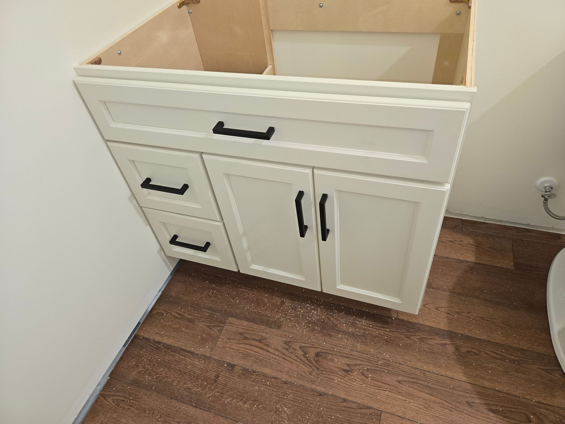 A white bathroom vanity cabinet with black handles, featuring a top drawer, two side drawers, and two cabinet doors.