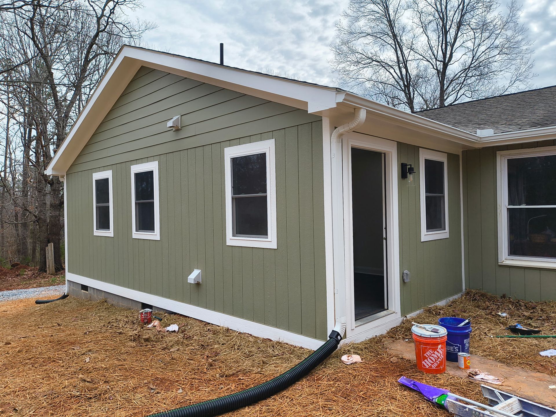 An exterior view of a green-sided house with white trim, multiple windows, a door, and a black downspout on a leaf-filled yard.