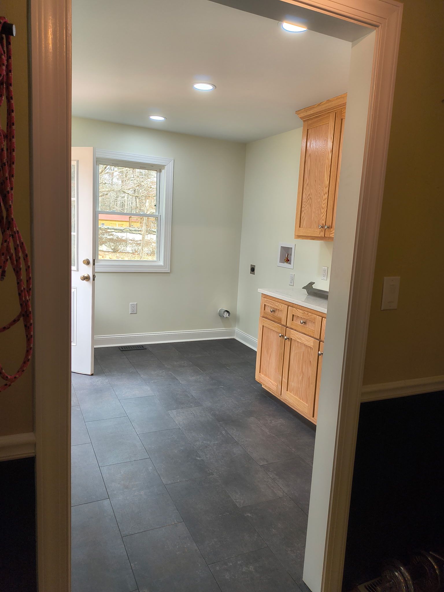 A view through a doorway into a small room with light gray walls, dark tiled floors, light wood cabinets, and a window.