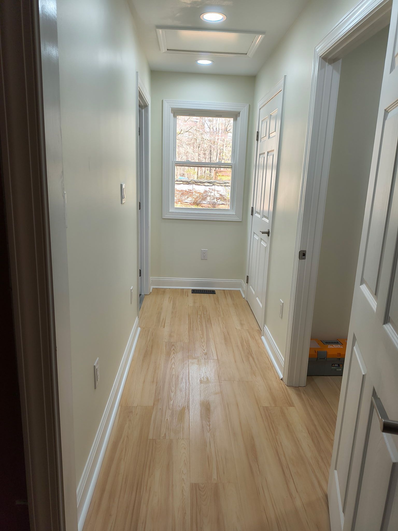 A narrow hallway with light wood flooring, beige walls, white doors, and a window at the end showing autumn trees.