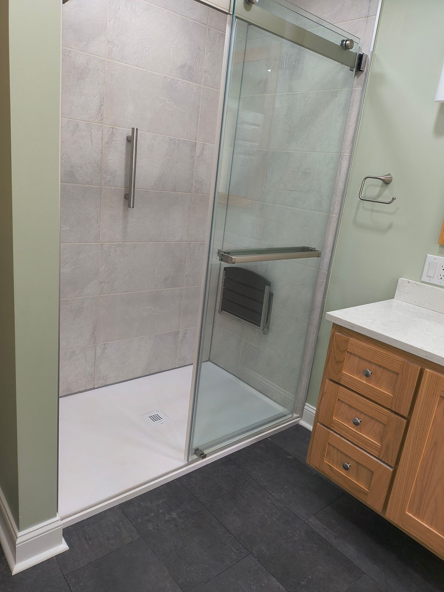 A walk-in shower with light grey tiled walls, a glass door, a mounted grab bar, and a foldable seat next to a wood vanity.