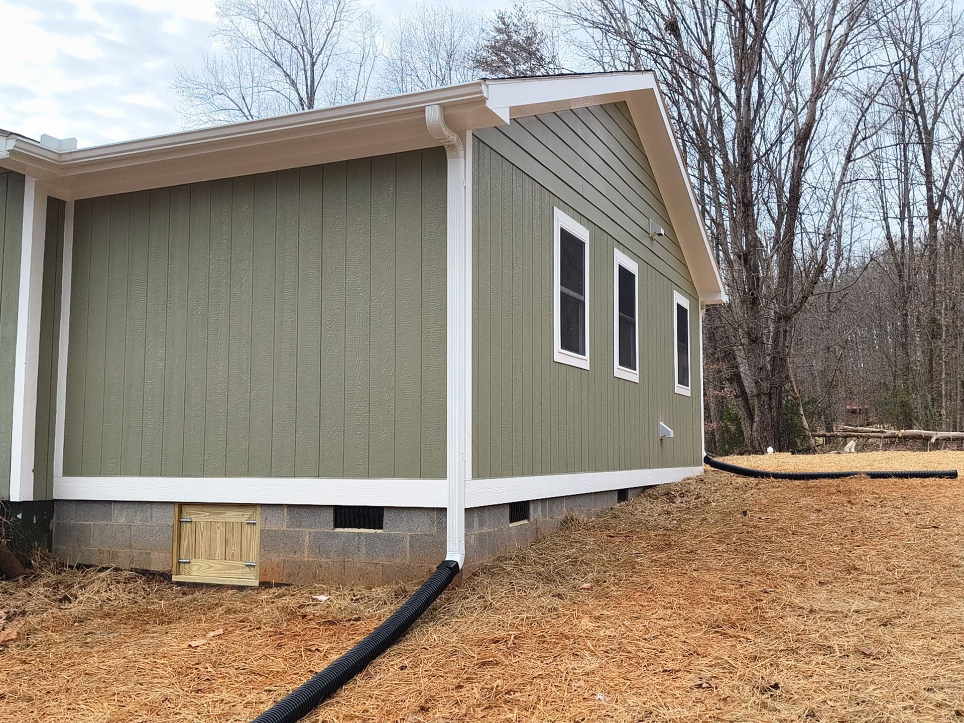 A corner view of a house with olive green board-and-batten siding, white trim, and a concrete block foundation.