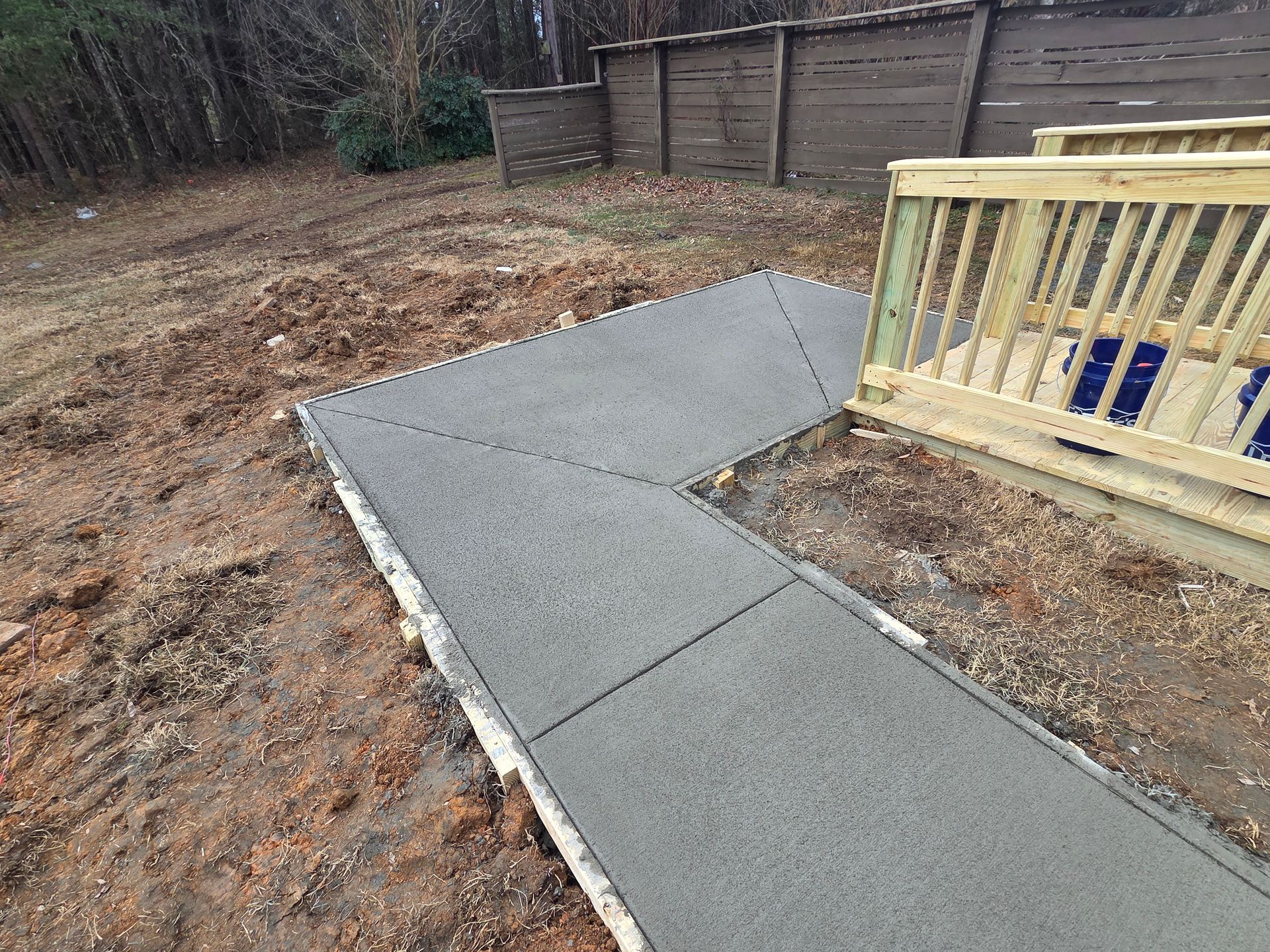 A newly poured L-shaped concrete sidewalk leading to a wooden deck in a yard with dirt and trees in the background.