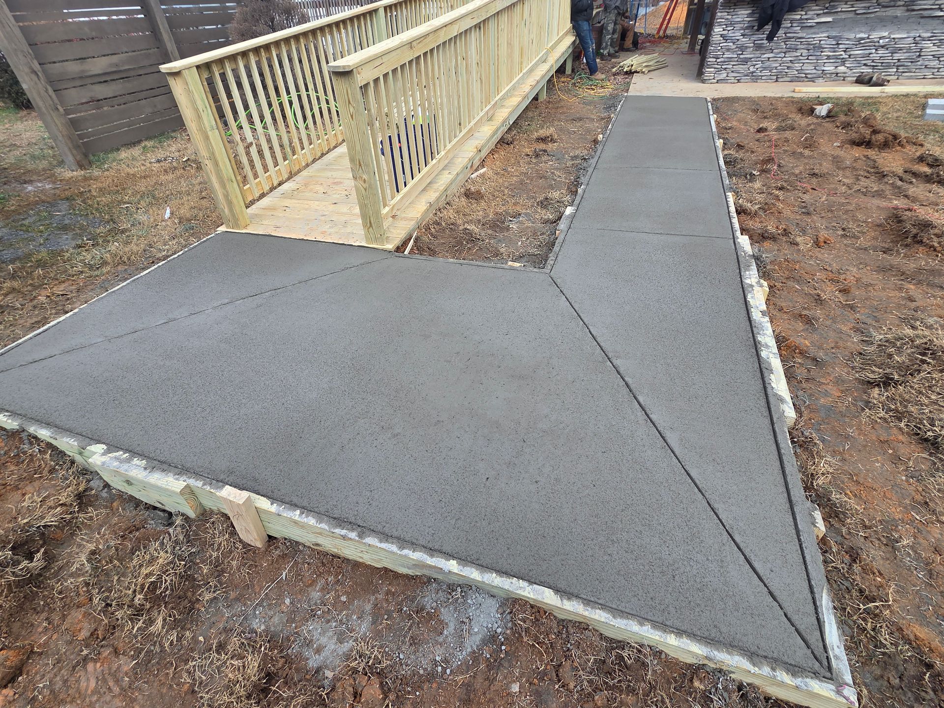 A newly poured concrete walkway connects to a wooden ramp under construction in a dirt-filled outdoor area.