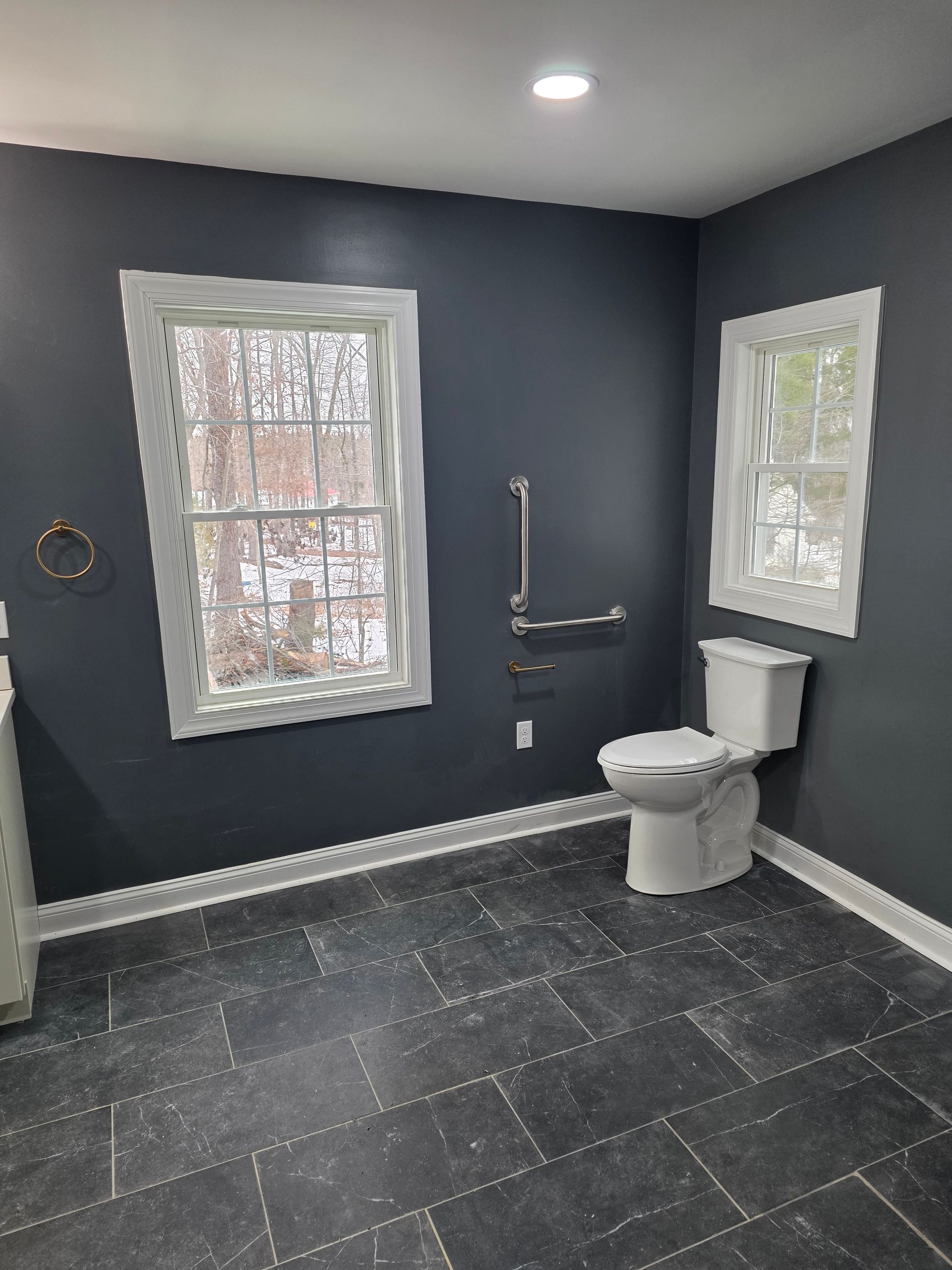 A bathroom with charcoal walls, dark stone-look tile flooring, two windows, and a toilet with a silver grab bar nearby.