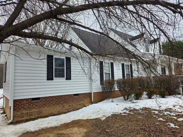 A white house with a brick foundation and black shutters, partially obscured by tree branches during winter.