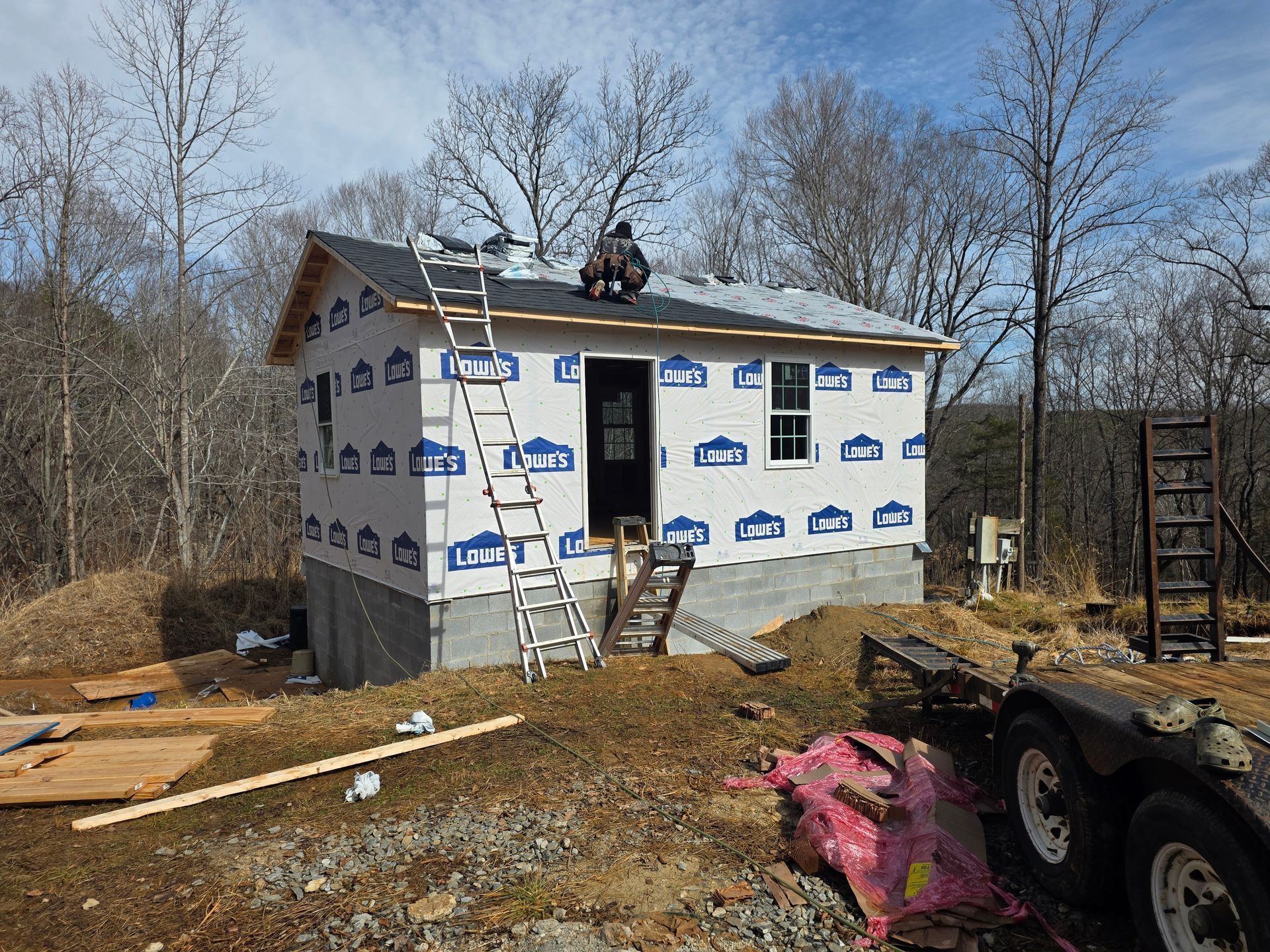 A person installing shingles on the roof of a small building under construction in a wooded area.