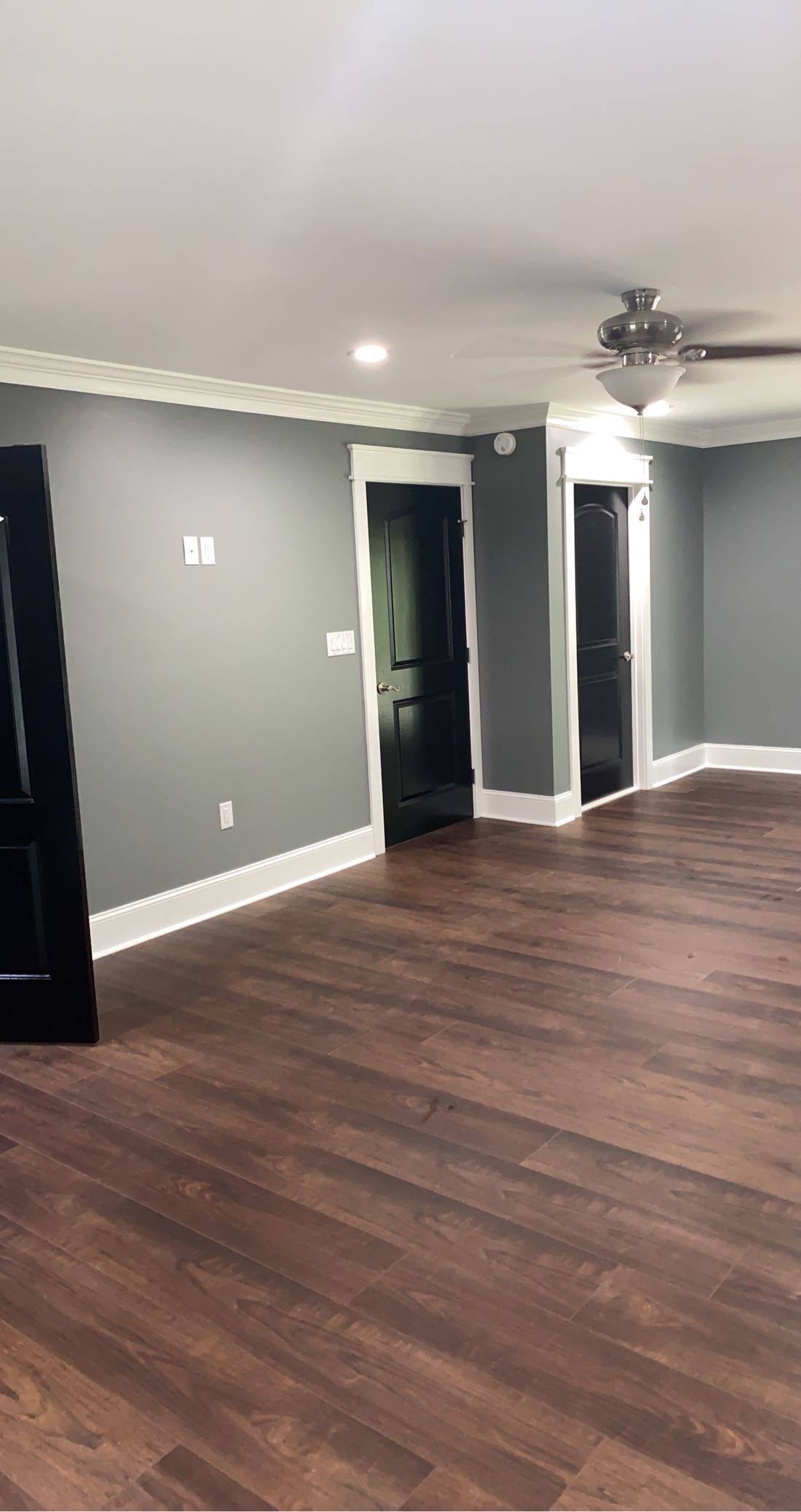 A room with grey walls, white trim, dark wood-look flooring, and three black doors under a ceiling fan.