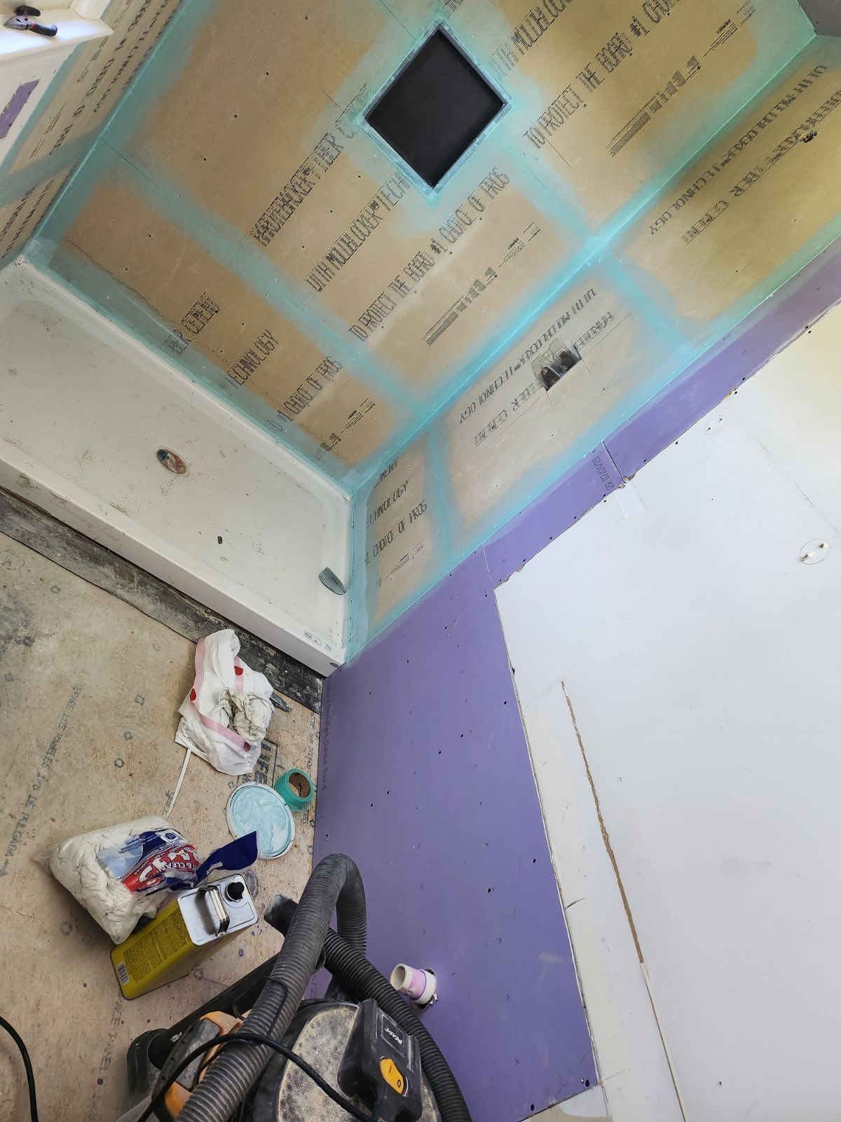 A shower stall under construction with light-colored tile backer board and purple drywall, showing floor debris and tools.