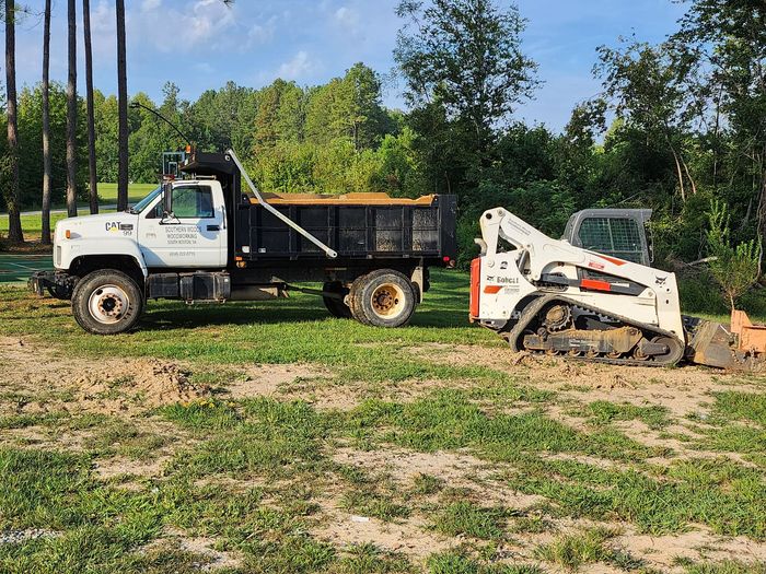A white dump truck parked next to a white and orange Bobcat skid-steer loader on a grassy construction site.