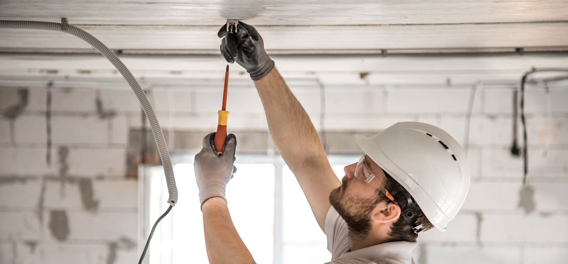 A construction worker in a hard hat and safety glasses installs electrical wiring into a concrete ceiling.