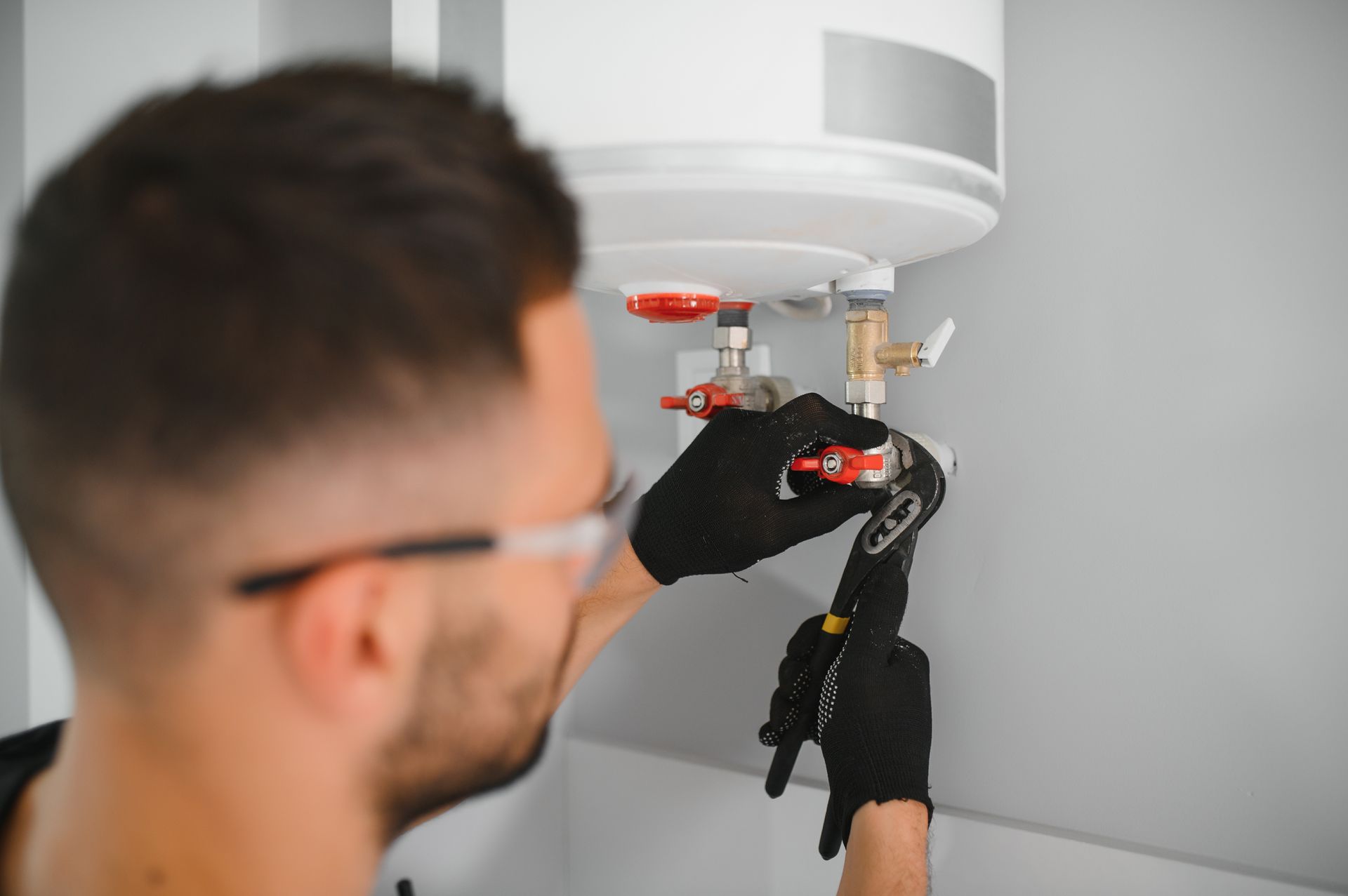 A person wearing black gloves and safety glasses uses a wrench to tighten a valve on a water heater.