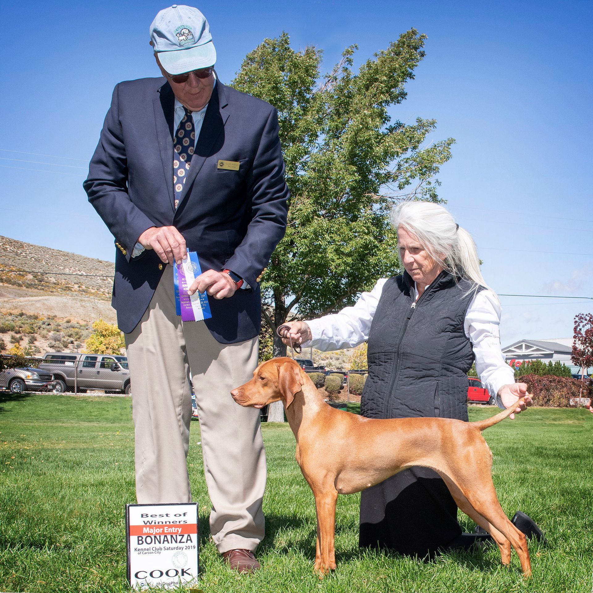 A man and a woman kneeling next to a dog holding a ribbon