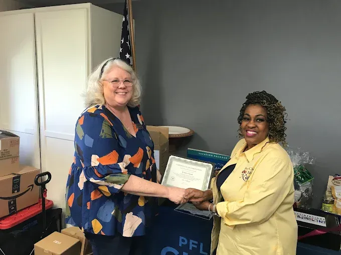 Two women, one in blue patterned top, one in yellow, holding a certificate indoors, smiling.