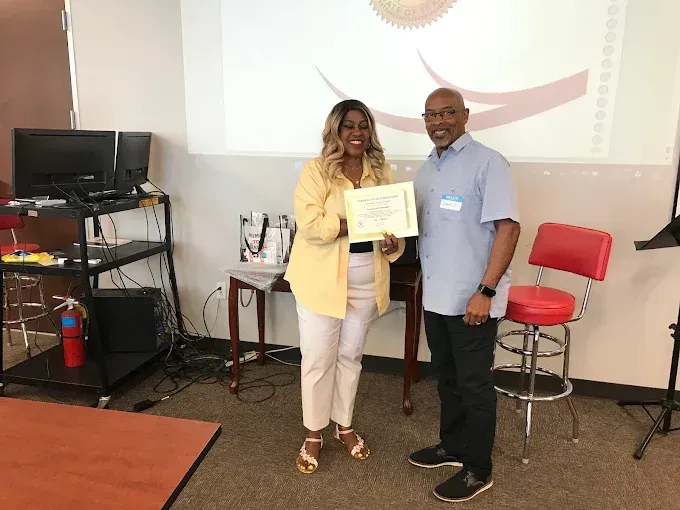Woman in yellow shirt and man in blue shirt hold a certificate; presentation in room.