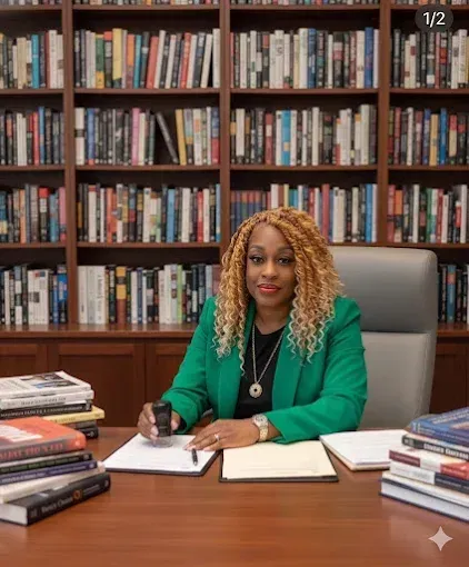 Woman in green blazer sits at desk in a library, surrounded by books, holding a stamp.