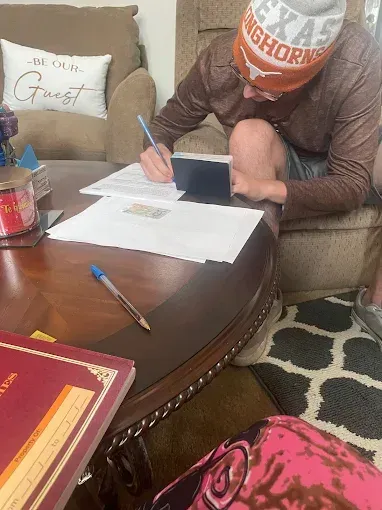Person wearing a Texas Longhorns hat, writing on paperwork at a coffee table with a book, pen, and phone.