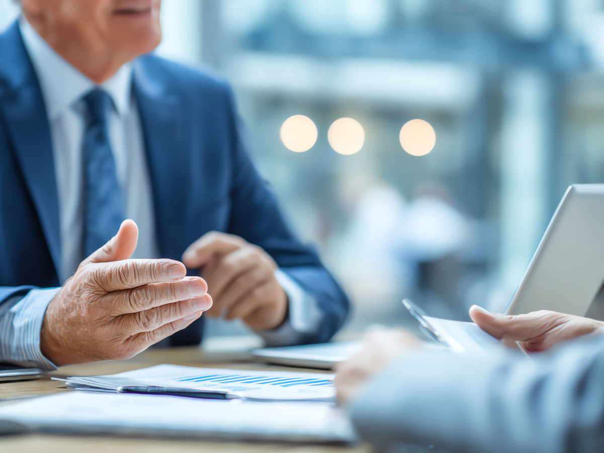 Two professionals in suits discuss documents at a table with a laptop in a brightly lit office setting.