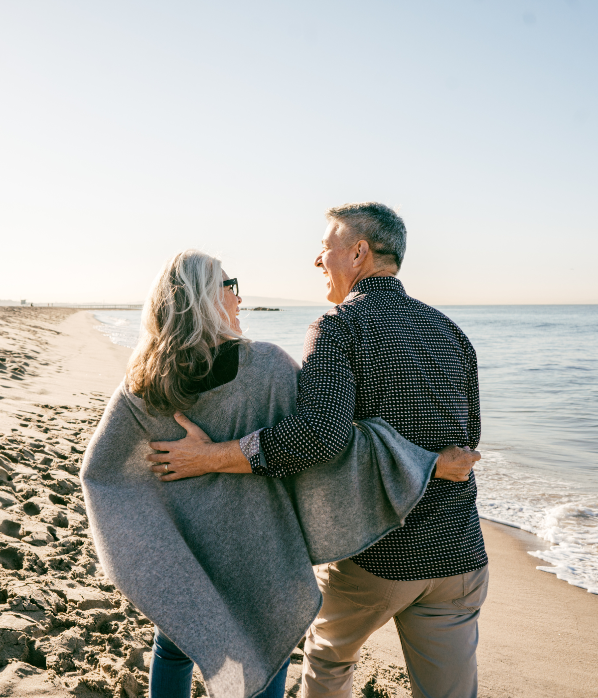 Couple embracing on a beach, looking at each other, laughing. Sunlight, ocean, sand.