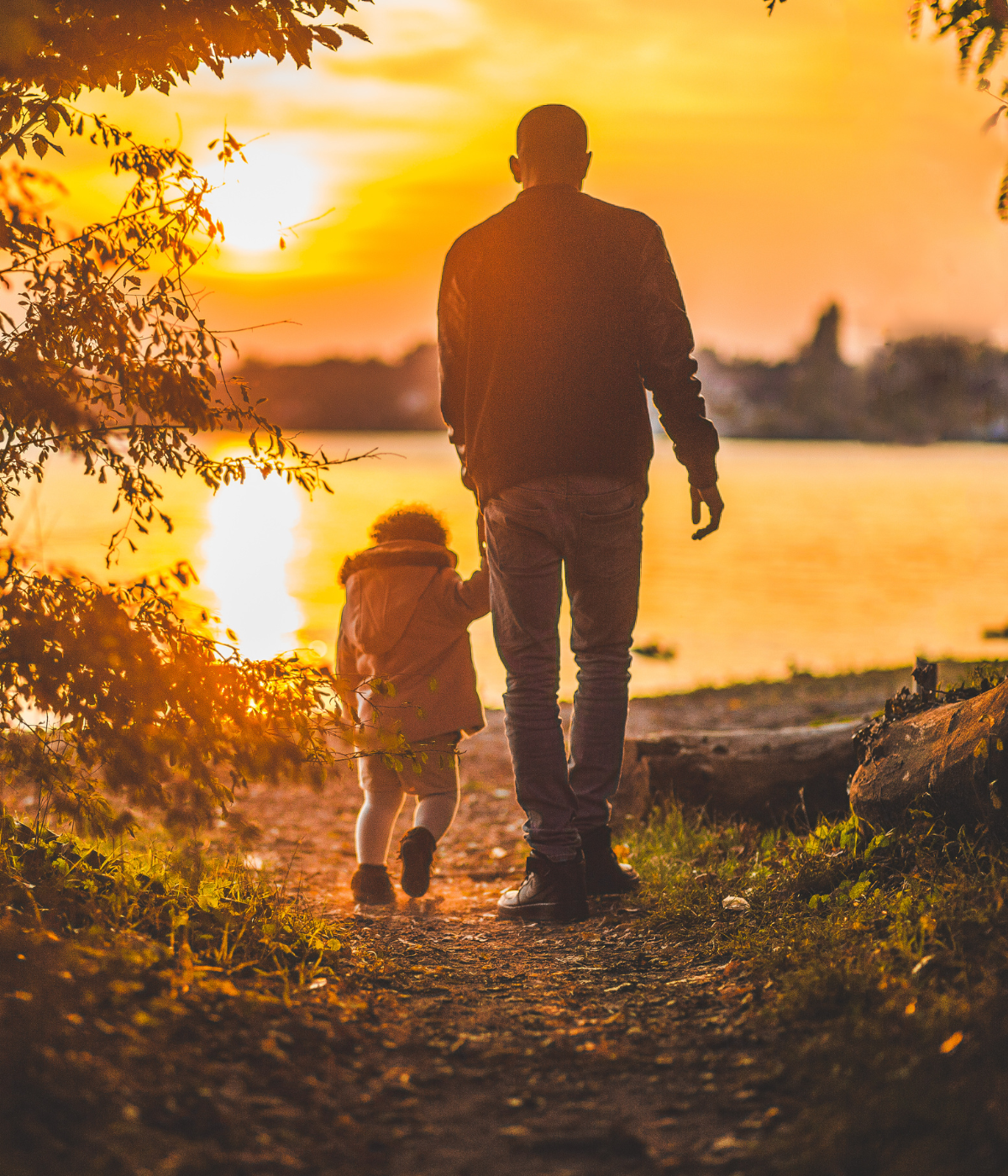 Father and child walking hand-in-hand along a path by a lake at sunset.