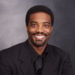 Professional headshot of a smiling person wearing a dark suit and shirt against a mottled gray background.