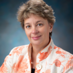 A person with short, light brown hair smiling at the camera, wearing an orange and white floral-print jacket.