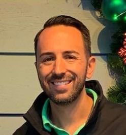 A person with a beard smiling against a light-colored wall with holiday greenery in the background.
