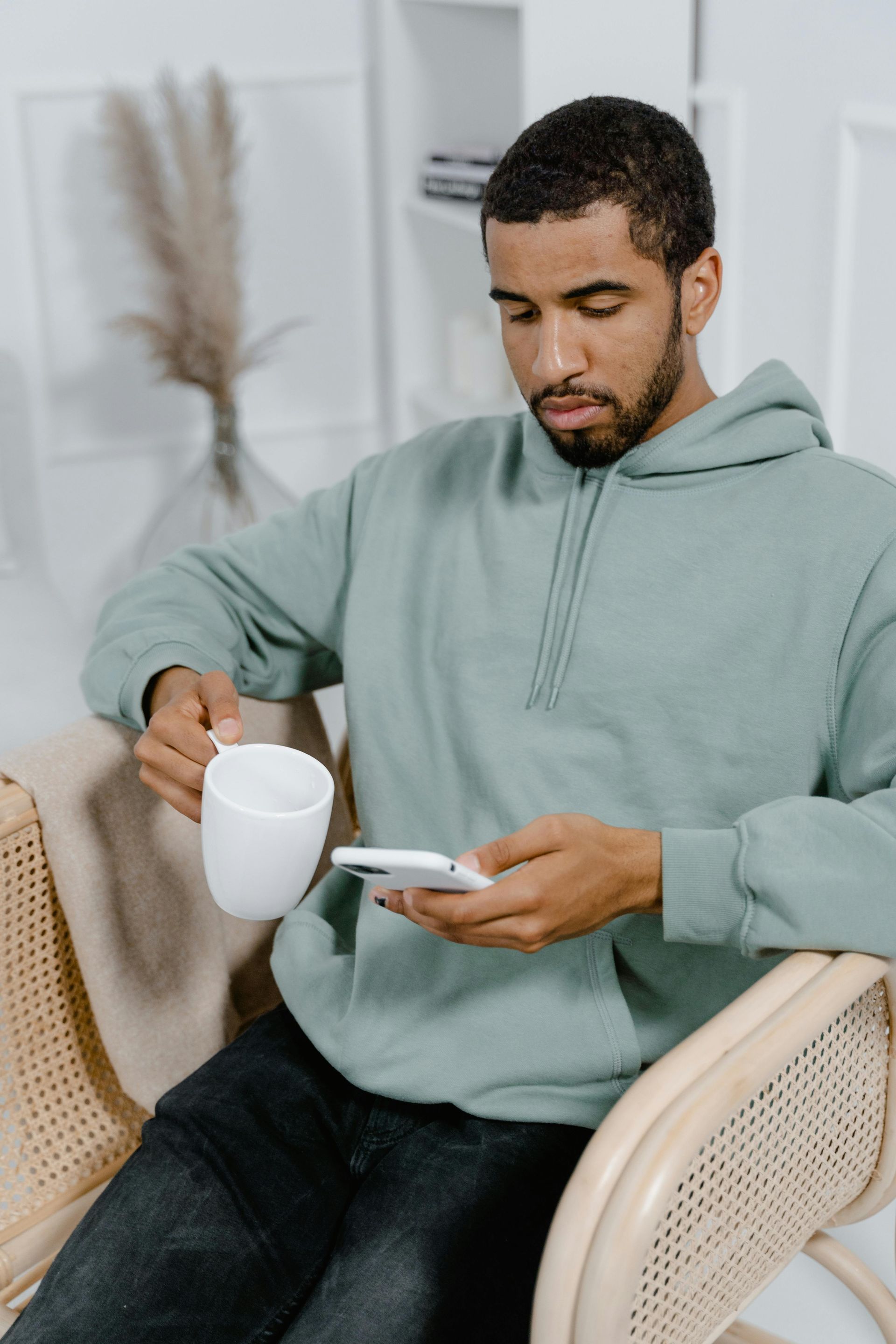 A person in a light green hoodie sits in a rattan chair, holding a white mug while looking at a smartphone.
