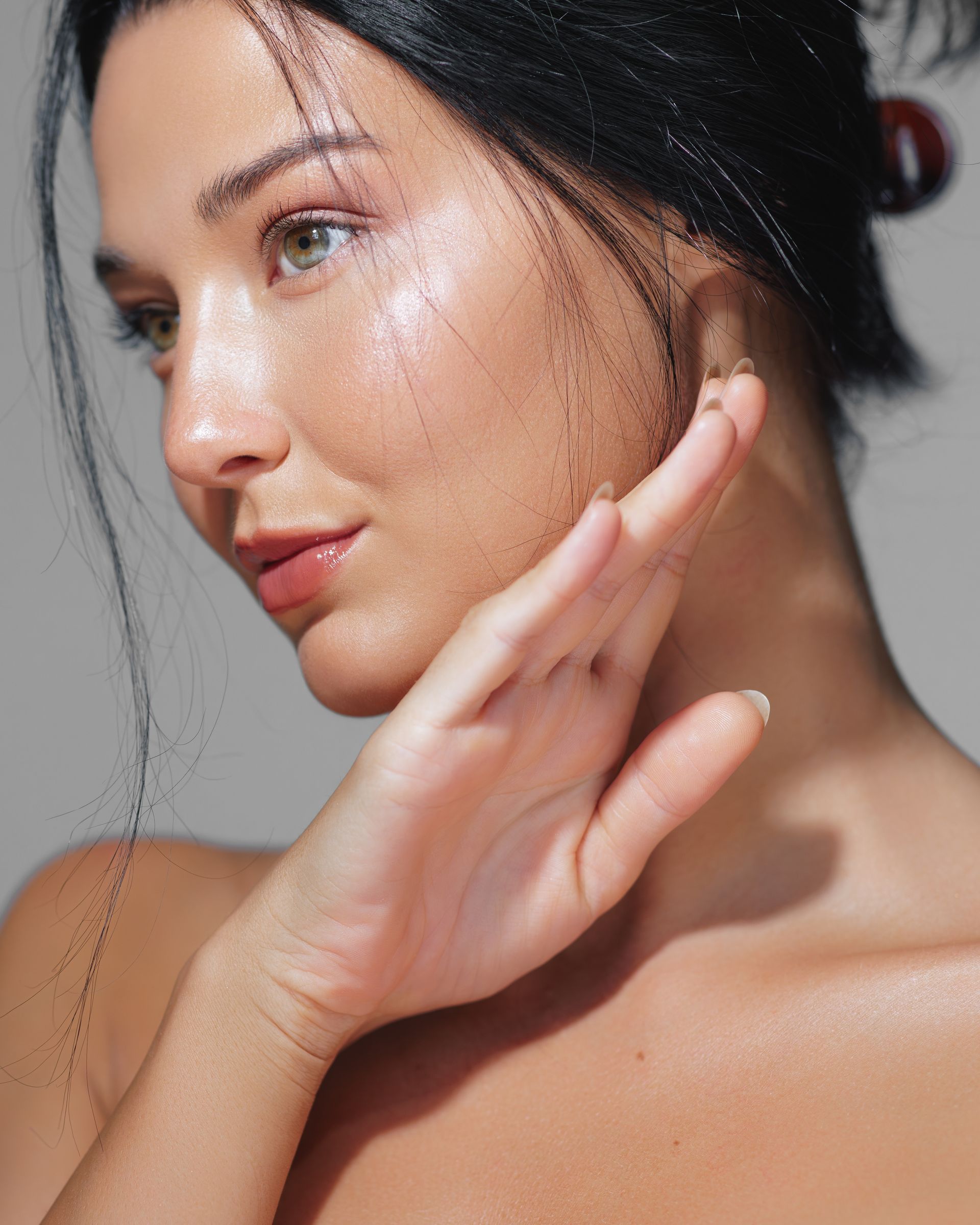 Woman with dark hair touches her cheek with open hand, slightly angled, against a neutral backdrop.