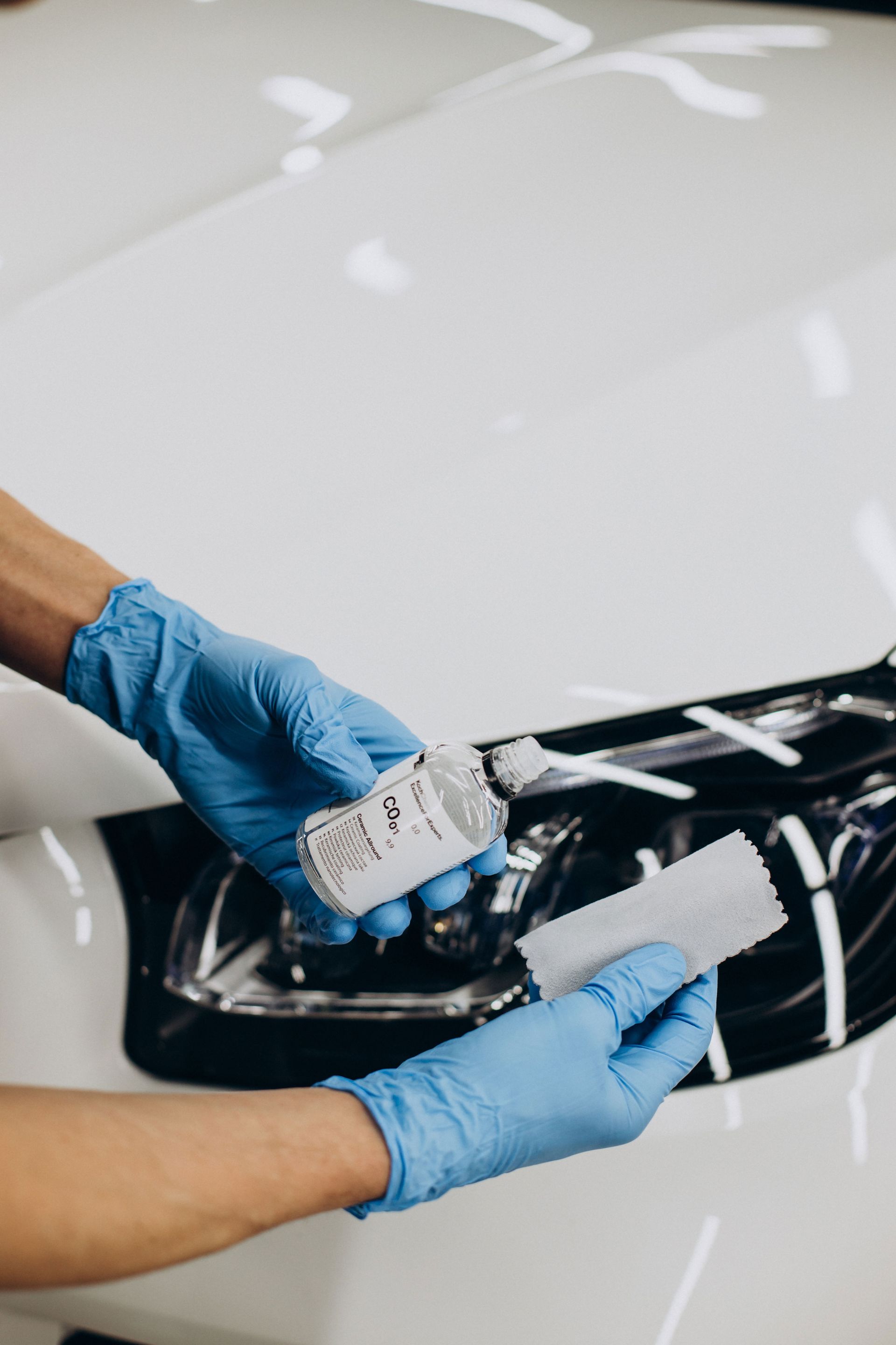 Person in blue gloves applying car wax to a white car using a sponge.