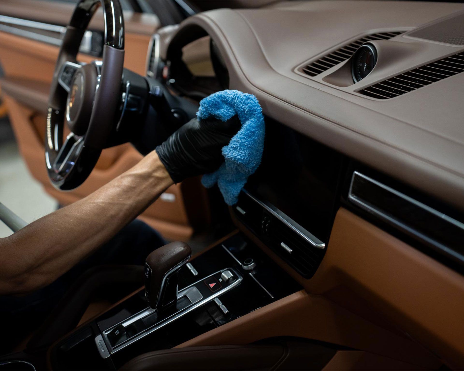 Person cleaning a luxury car's interior with a blue cloth. Brown leather dashboard and black gloves visible.