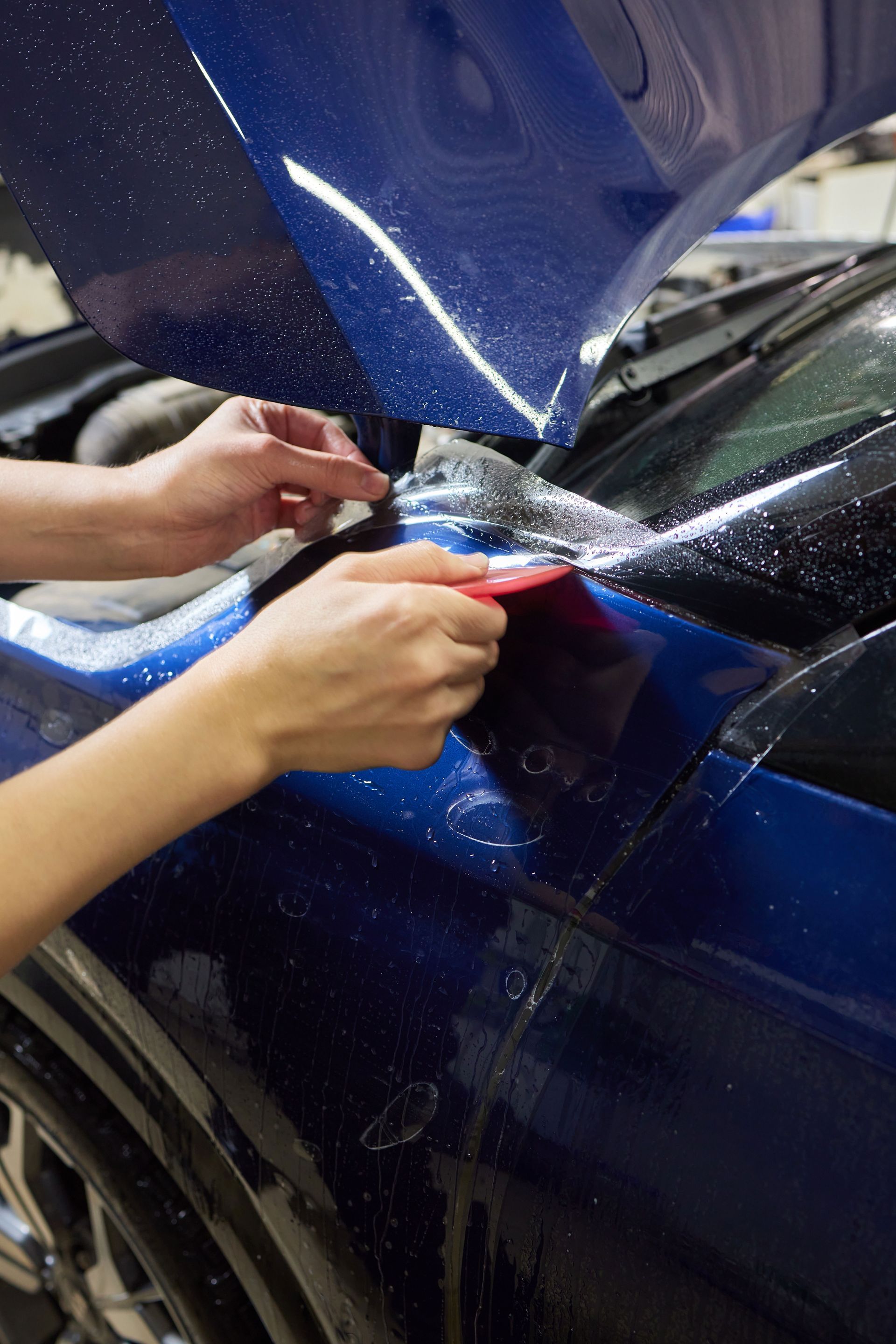 Person applying clear protective film to a blue car's hood using a squeegee.
