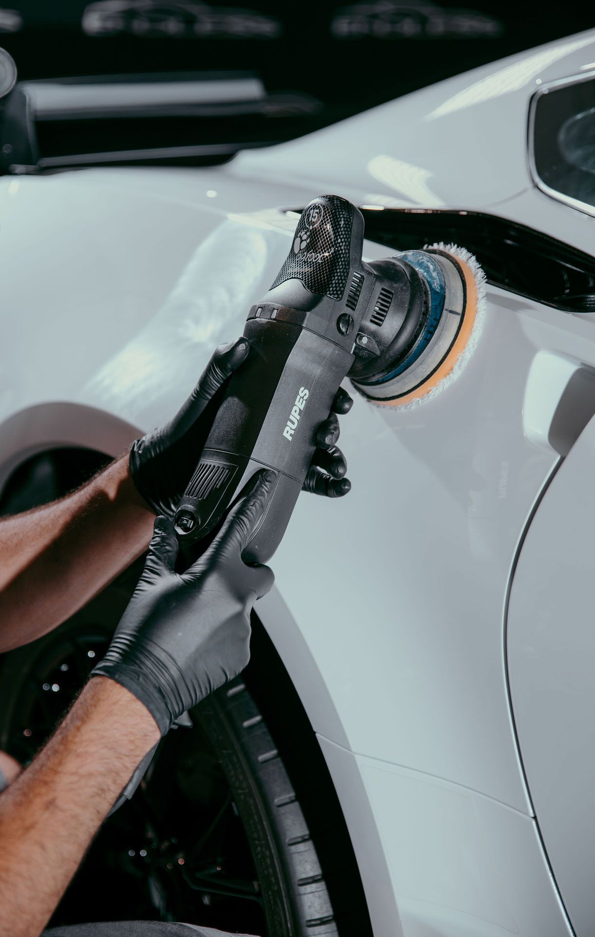 Person polishing a white car's side panel with a power buffer while wearing black gloves.