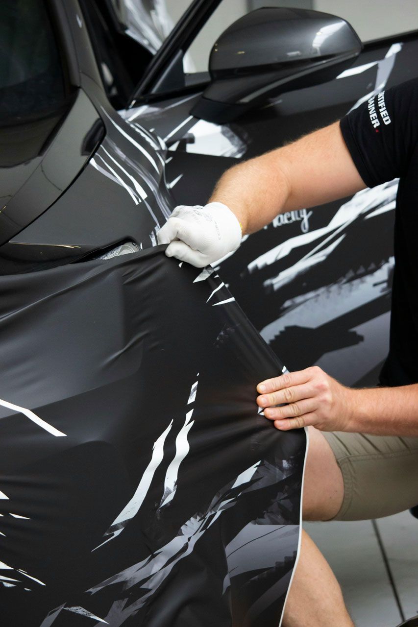 Person applying black vinyl wrap to a dark gray car, wearing gloves.