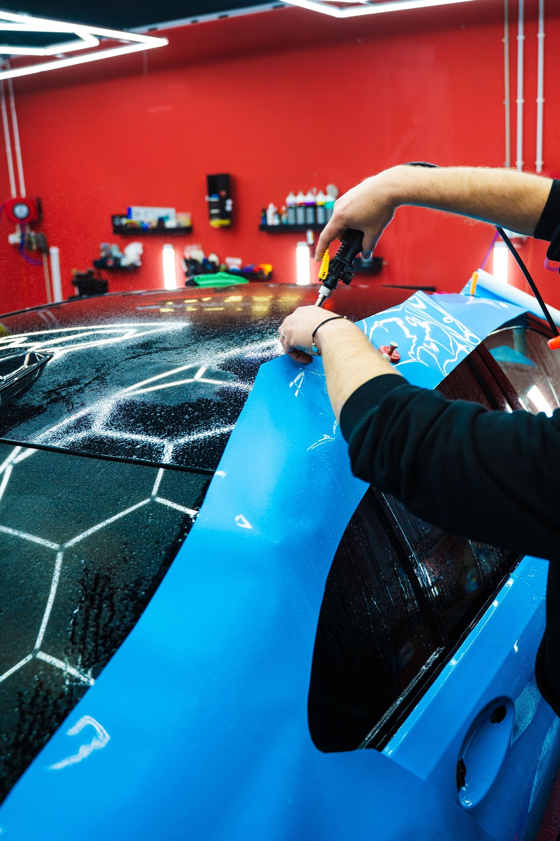 Person applying blue vinyl wrap to a car's window and body panel in a shop with red walls.