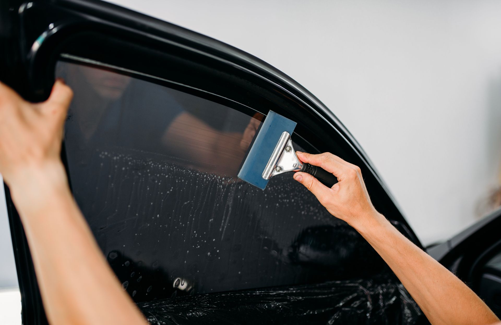 Person applying tint to a car window with a blue squeegee. Dark car door, white background.