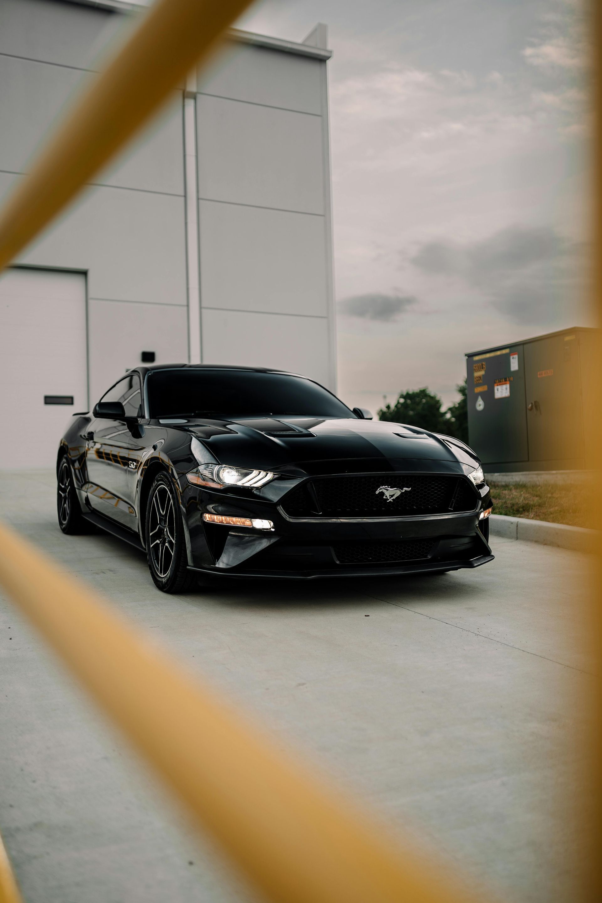 Black Mustang coupe parked in front of a white building, seen through yellow metal beams.