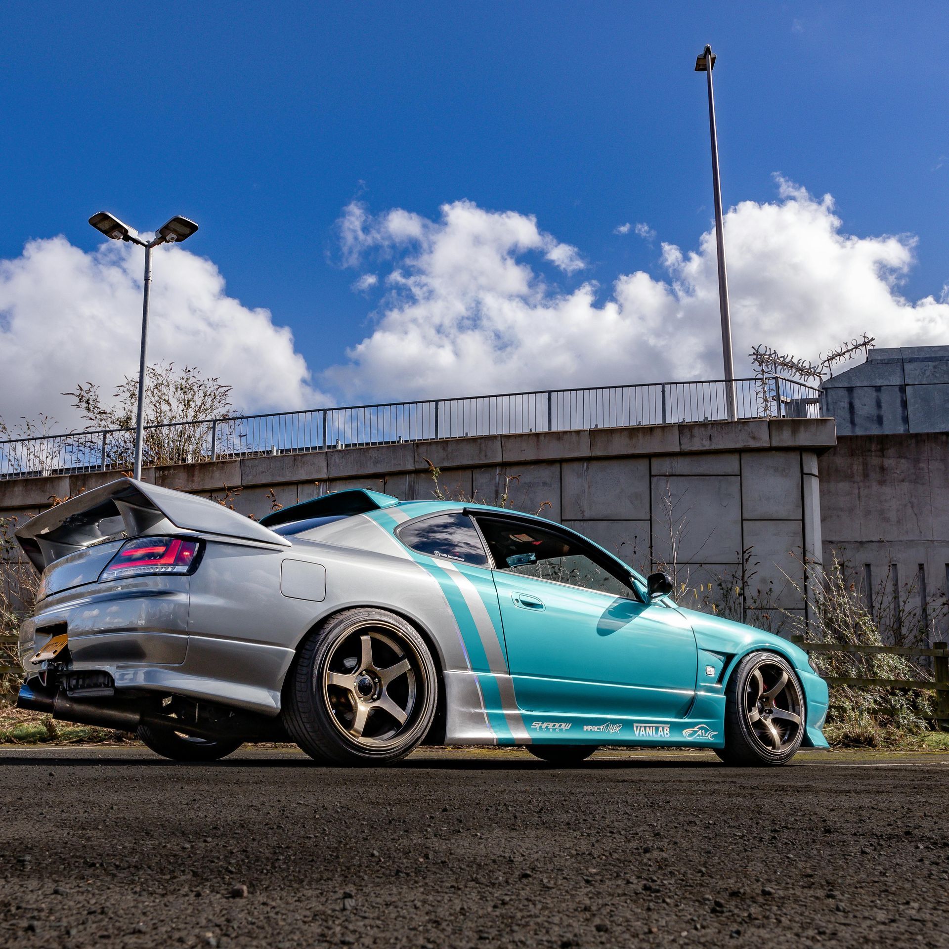 Silver and turquoise modified sports car parked near a concrete wall under a blue sky.