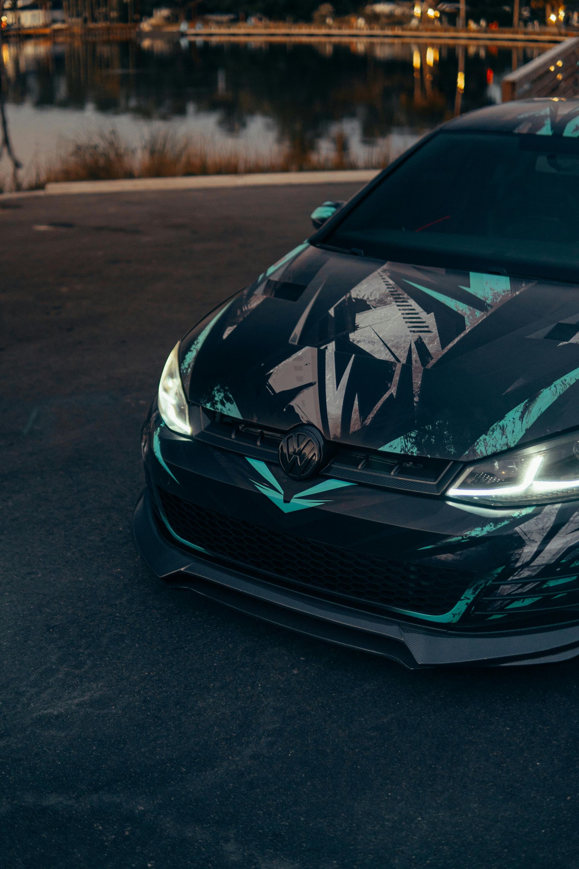 Black car with teal accents parked by water at dusk.