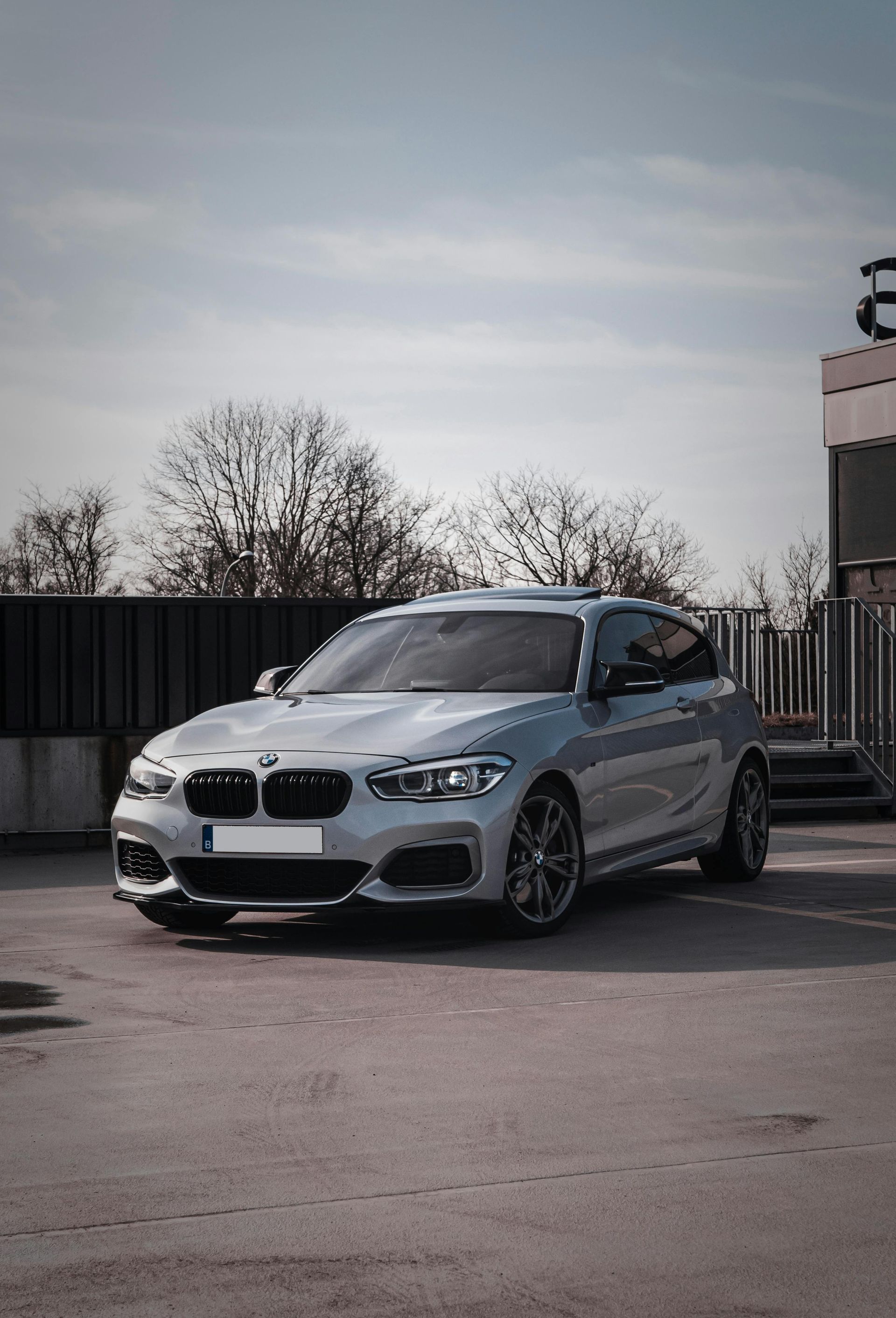 Silver BMW coupe parked on a rooftop in front of a gray fence and bare trees under a cloudy sky.