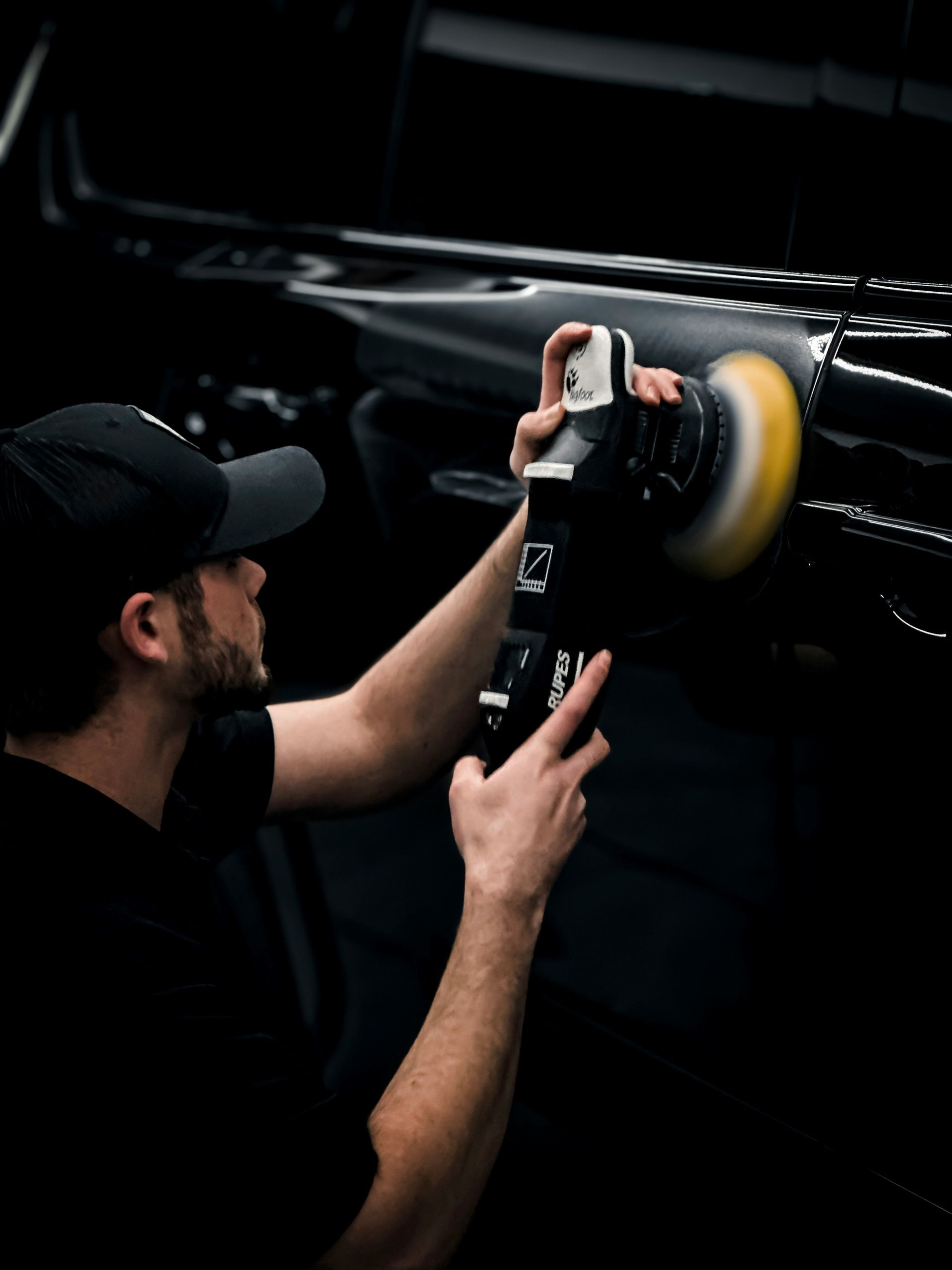 Man polishing a black car with a rotary buffer, indoors.