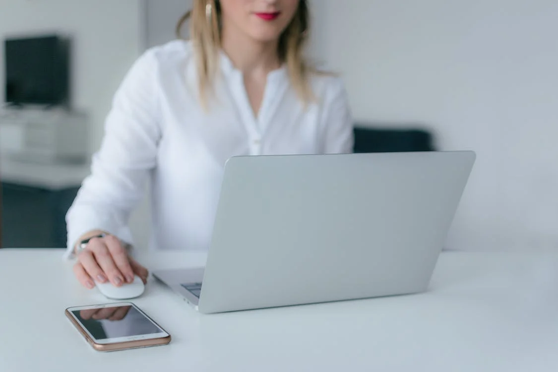 Femme en chemise blanche utilisant un ordinateur portable à un bureau, avec une souris et un téléphone.