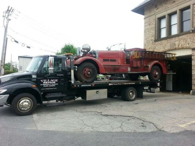 Tow truck carrying a vintage red car parked beside a brick building | Village Repair